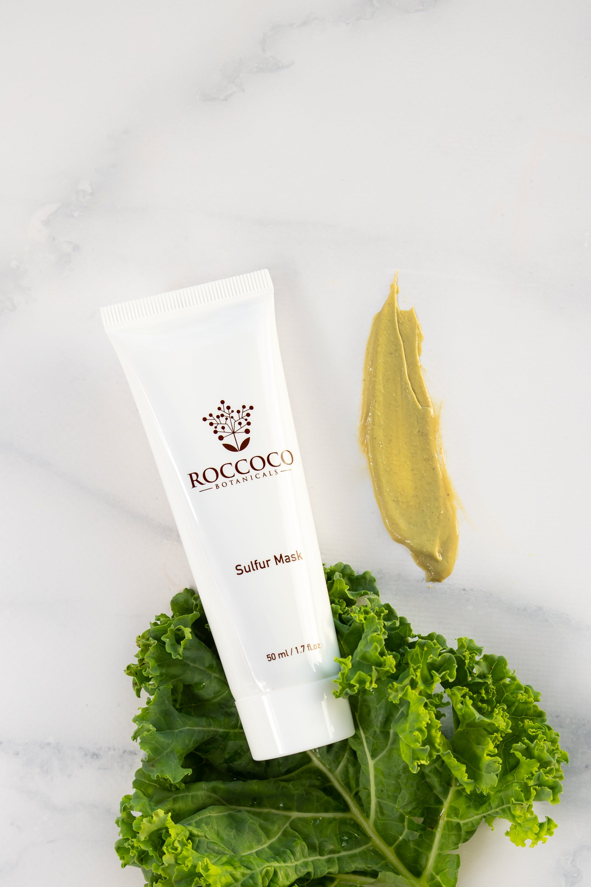 Sulfur Mask tube against a kale leaf with dab of the golden yellow mask on the side